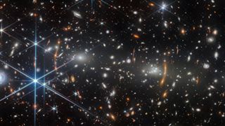 A black space background is speckled by yellow, white, blue and orange stars including blue stars with long diffraction spikes.