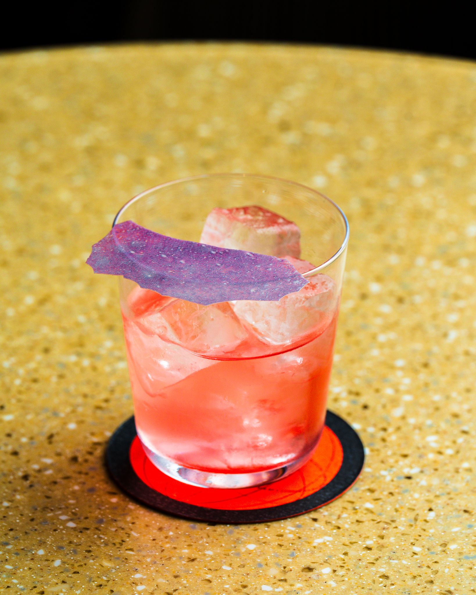 A grapefruit-shaded cocktail poured into a normal water glass and topped with a purple, iridescent, leaf-like crunchy topping sits on a terrazzo ochre and green table.