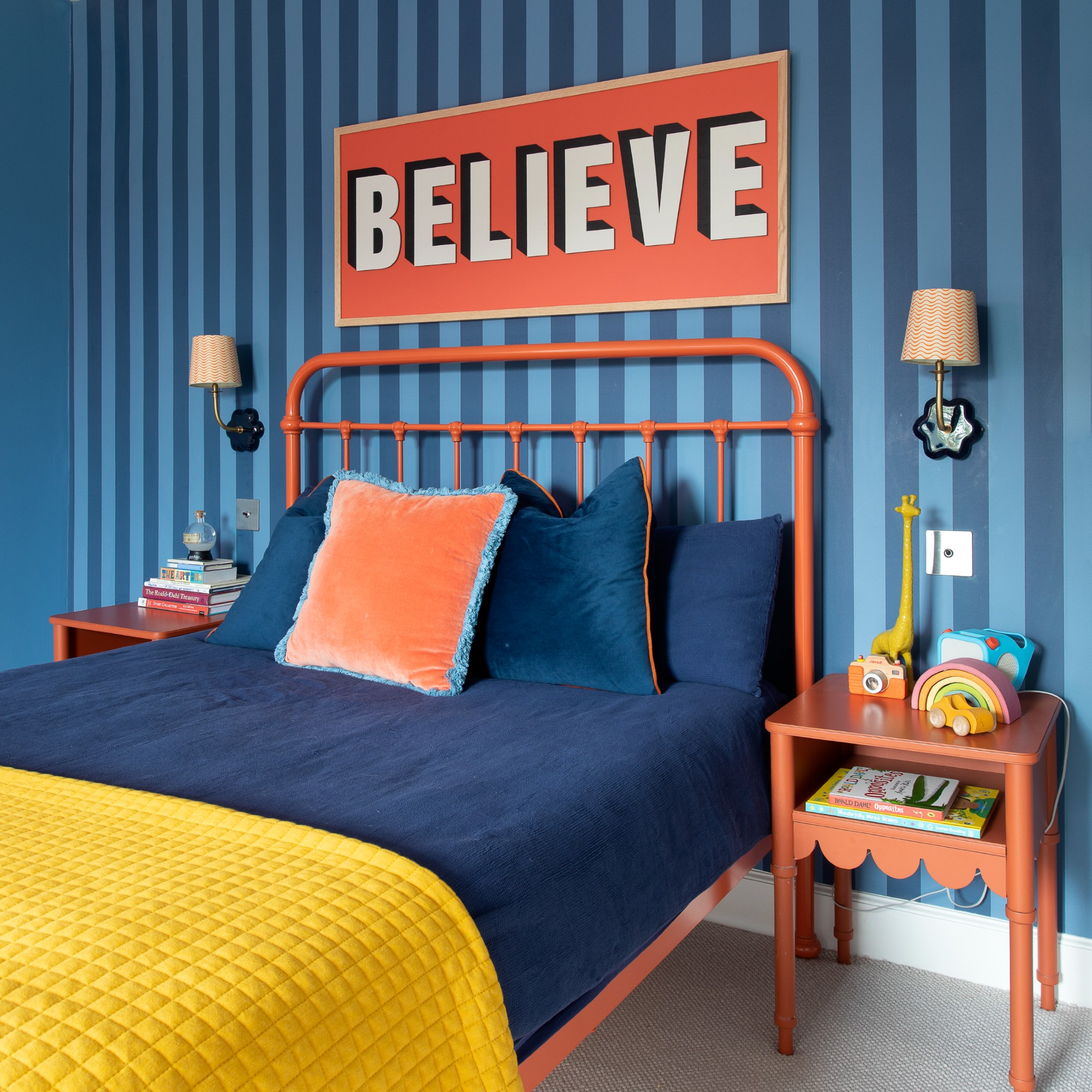 A blue bedroom with a striped feature wall and coral metal bed with matching bedside tables and wall sconces on either side of the bed