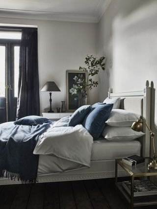 White bedroom with blue cushions by Neptune, gold metal and glass bedside, mirror, gray table lamp, dark gray drapes, blue blanket and cushions