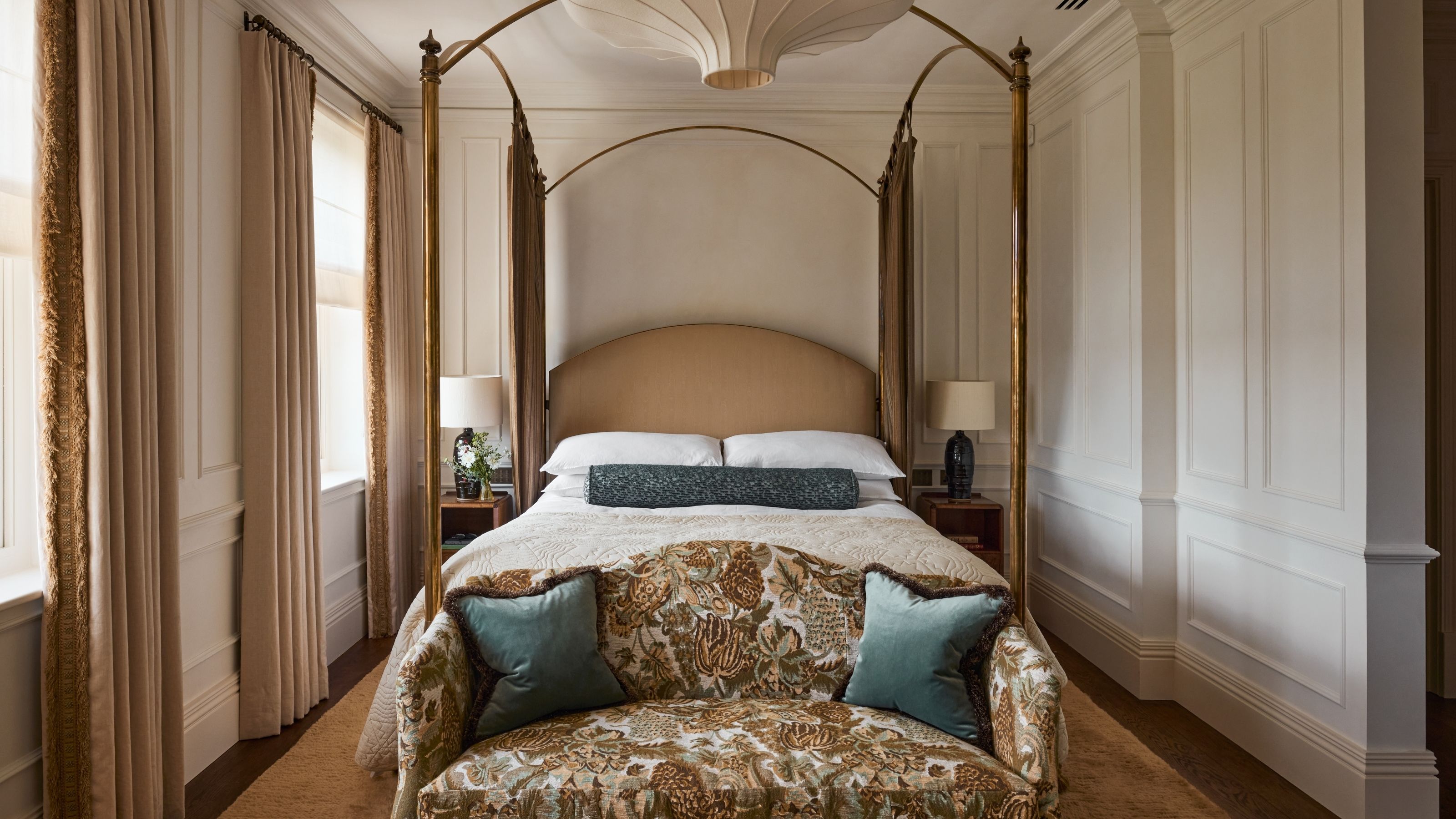 a soft neutral primary bedroom with paneled walls, four poster brass bed, silk pendant nad tapestry print end of bed settee