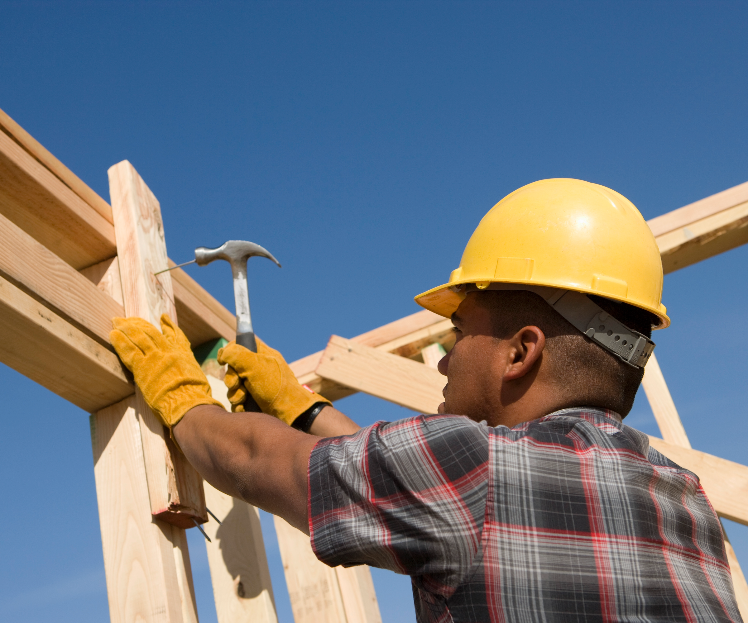 Person wearing a builder&#039;s hat and hitting a hammer into a nail on a wooden structure
