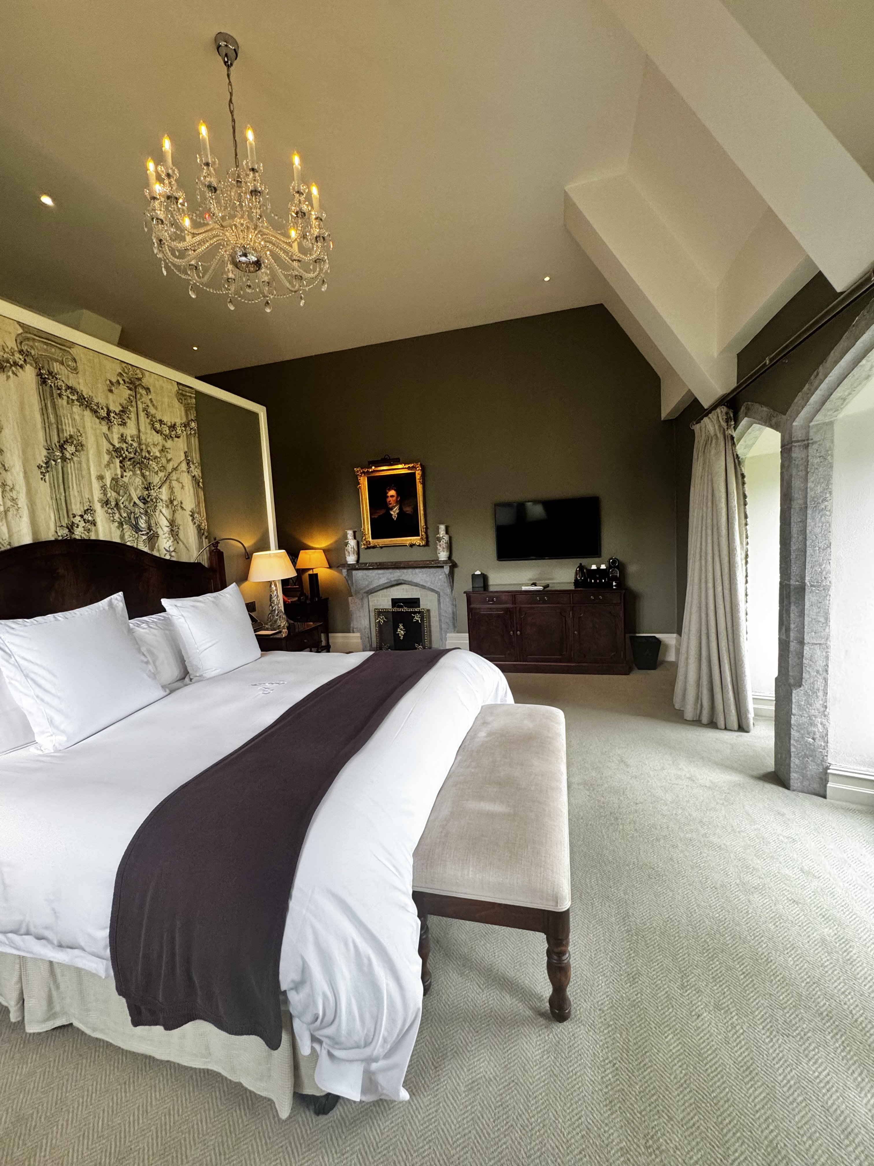 A hotel room with a large bed and a chandelier with windows surrounded by stone arches