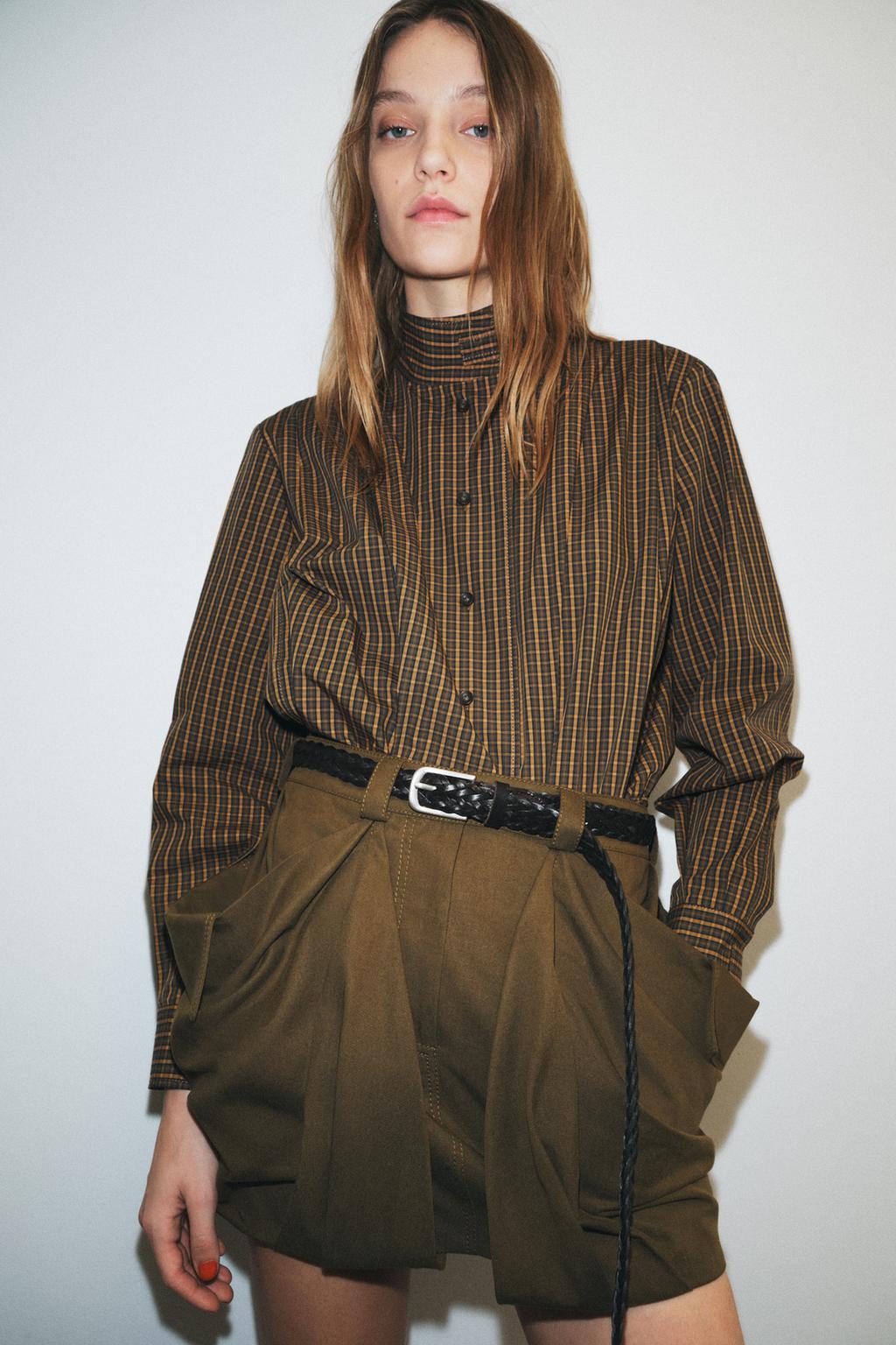 Model wears plaid long sleeve button down mock neck top and cargo shorts with black belt.