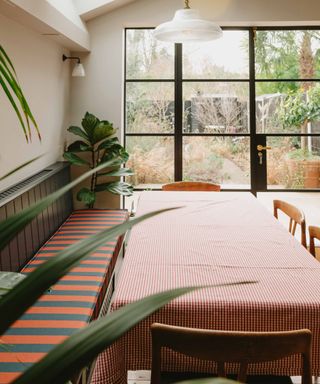 Stylish dining nook with a long built-in banquette in striped fabric, a large dining table with gingham cloth, and garden views framed by black steel-framed windows.