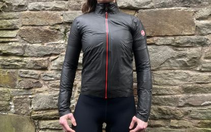 This image shows the torso and arms of a woman wearing the Castelli Idro 3 jacket in black. Behind them is a stone wall. 