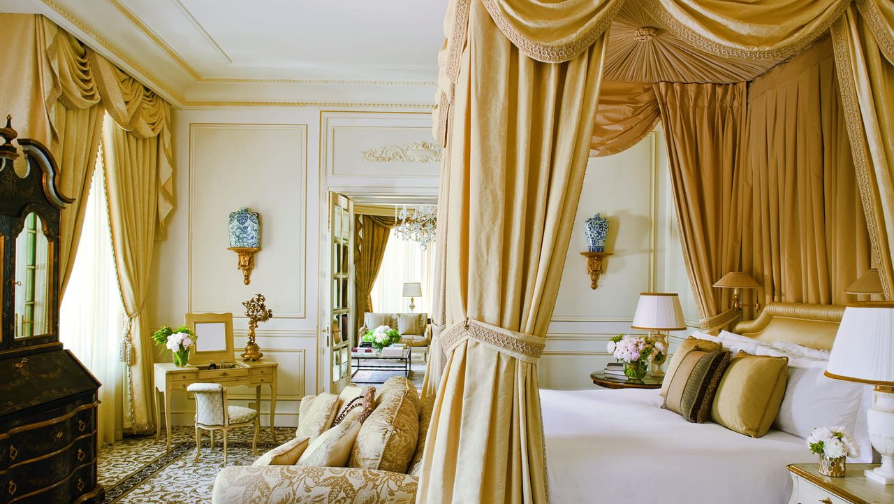 Traditional bedroom with white walls that have crown molding. There are luxurious gold curtains on the window and a matching set as the bed canopy.