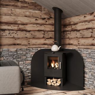 A modern wood-burning stove sits on a curved black hearth against walls of rustic wood paneling and stone cladding. A grey armchair and geometric rug complete the cosy space, illuminated by natural light from a large window.