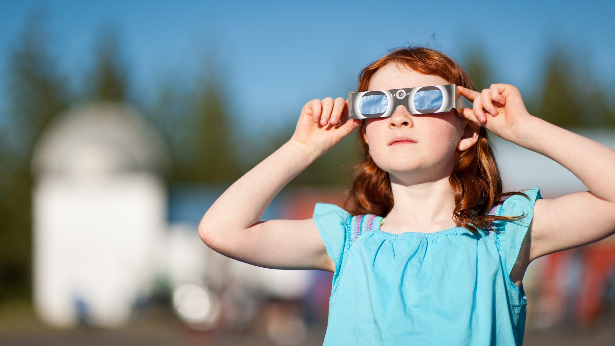 Una chica con una blusa azul sostiene un par de anteojos de eclipse en su cara mientras mira un eclipse.