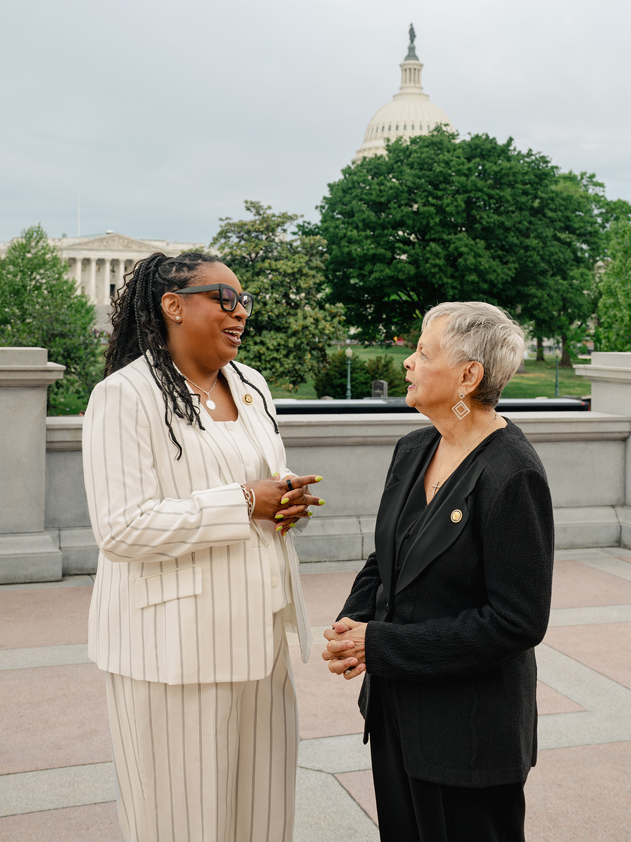 LaMonica McIver and Bonnie Watson Coleman face each other in conversation on the Capitol grounds, the Capitol dome visible behind them. McIver wears a white pinstripe suit; Watson Coleman wears black.
