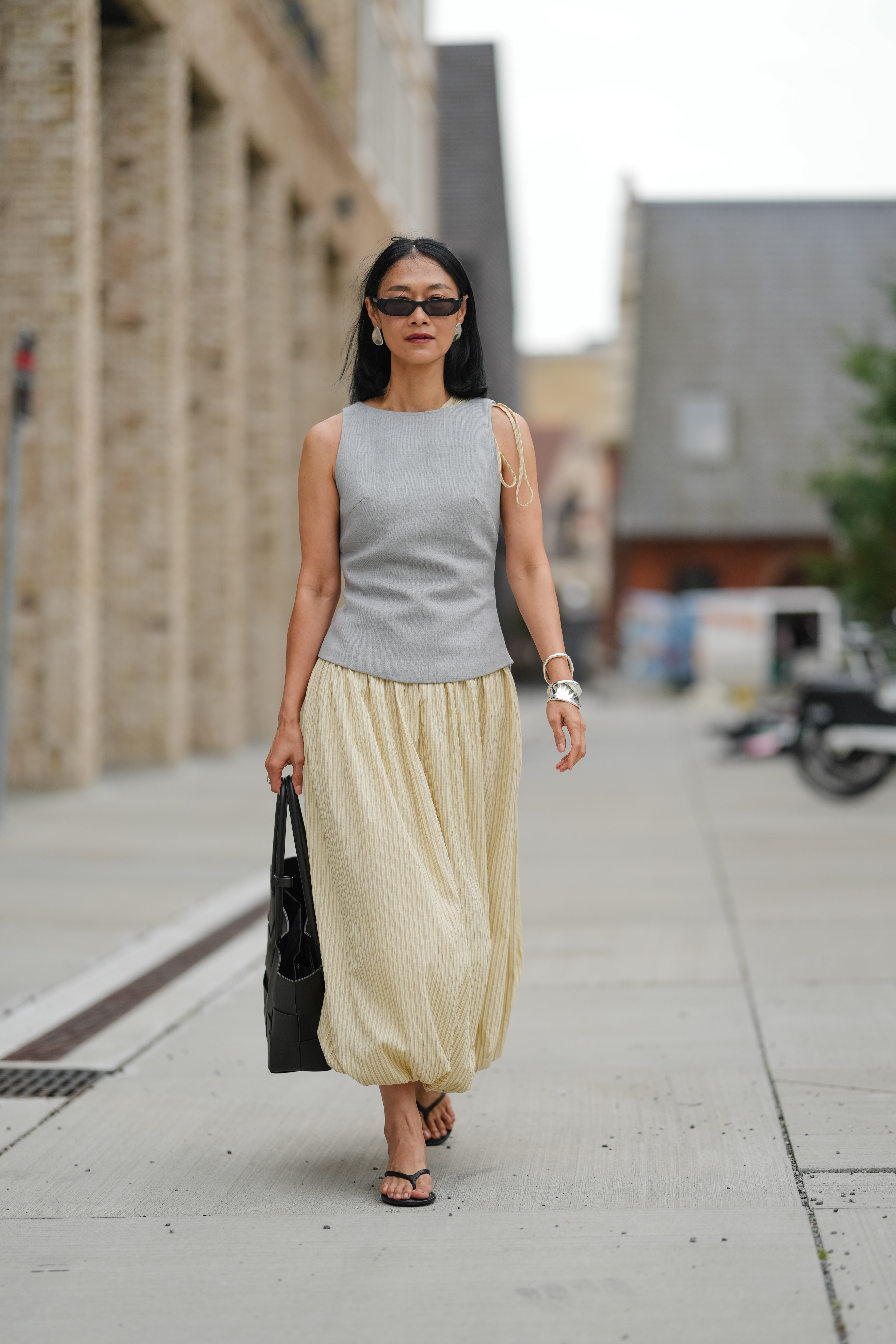 Pornwika Spiecker wears a sleeveless, light gray top with a round neckline and subtle side tie details. Black rectangular sunglasses cover the eyes. Hair is styled straight and loose. A large black woven tote bag is carried on the shoulder. The skirt is a voluminous, ankle-length piece in a light cream color with a subtle vertical stripe texture. Footwear consists of black thong sandals with flat soles, outside Birrot, during Copenhagen Fashion Week, on August 07, 2025 in Copenhagen