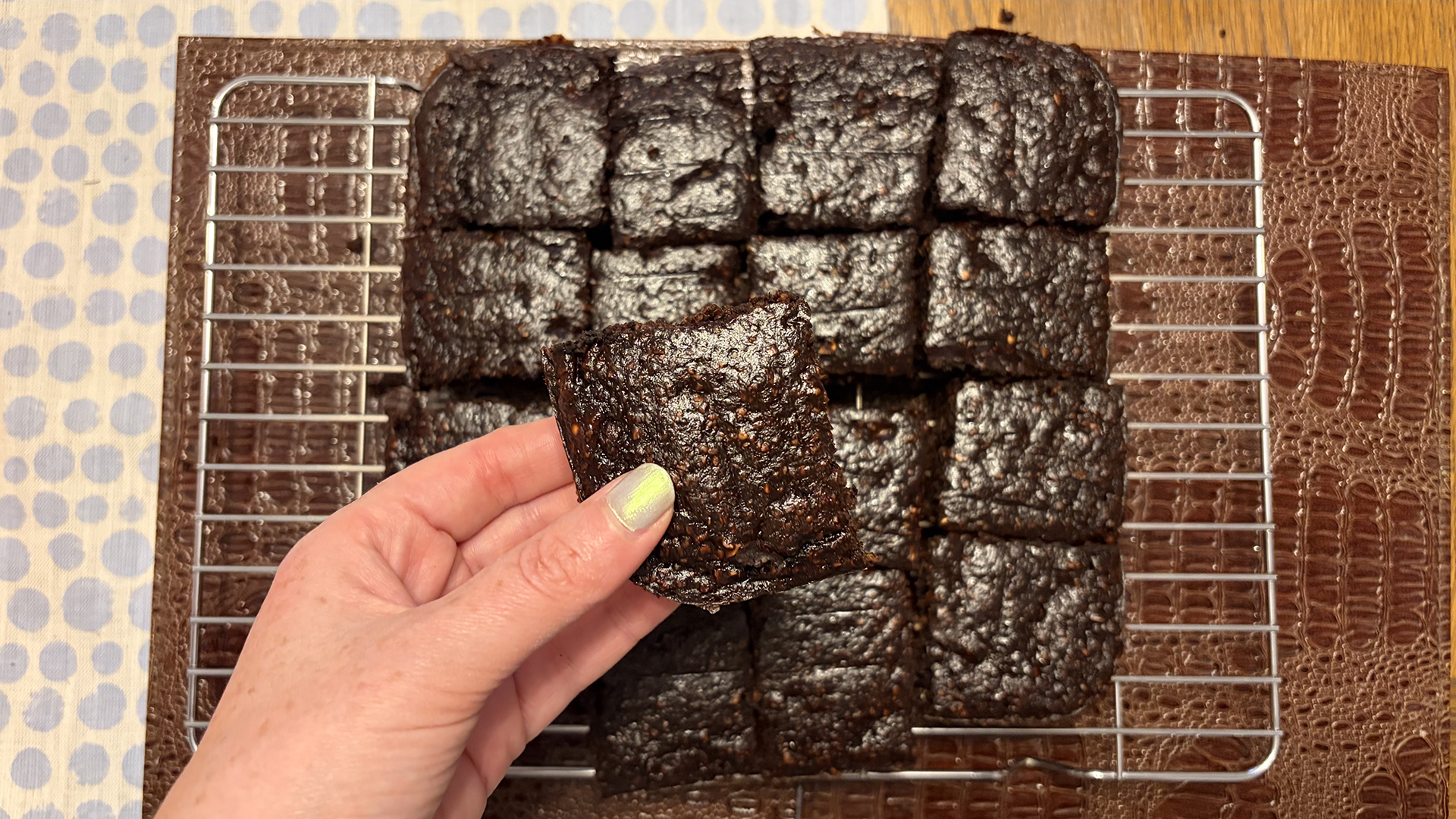 A hand holds a brownie to the camera. Beneath is is a pan of freshly backed brownies on a cooling rack