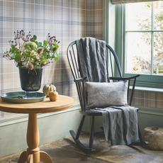 Grey rocking chair with grey throw and cushion by a sash window with tartan wallpaper on the wall.