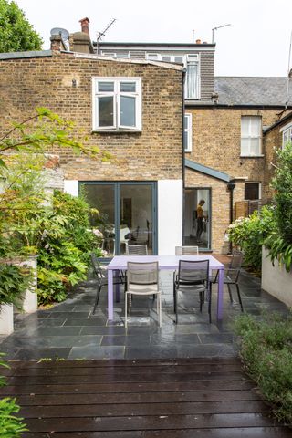 a side return single storey extension exterior, with a grey patio with a purple and black dining set on it, and a wooden deck in the foreground