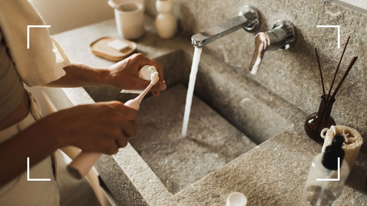 Woman&#039;s hands holding electric toothbrush under the tap, across stone sink with mirror above, demonstrating why are my teeth turning brown