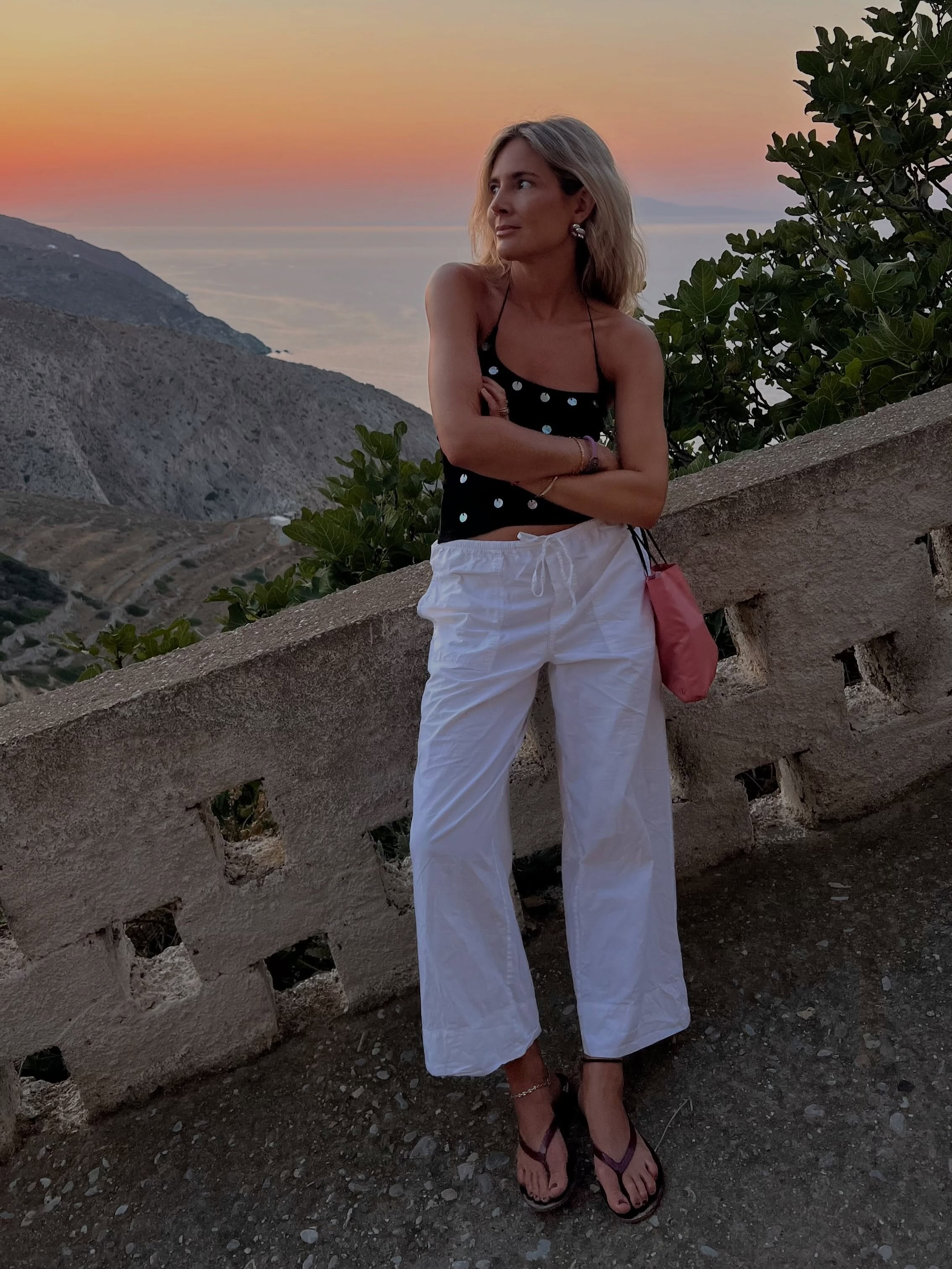 An image of an influencer in front a stone wall at sunset wearing a black embellished camisole, white cotton trousers and black flip-flops holding a pink handbag