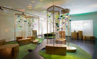 A lounge space consists of brass seating modules, with Plexiglas structures in different shapes and colours falling down from the ceiling. There is a green carpet below the seating modules.