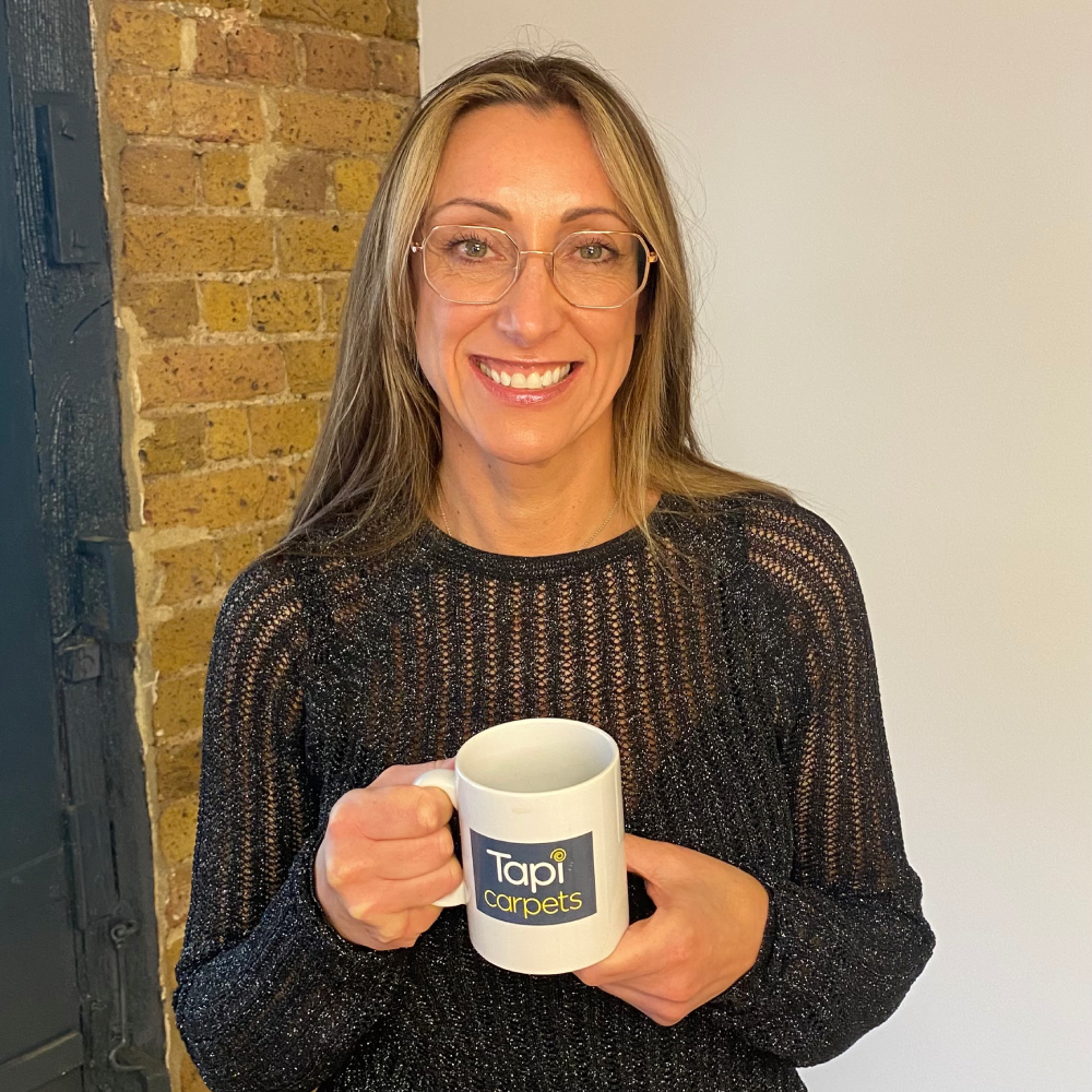 Female with long blond hair and metal framed glasses wearing black top and holding mug with Tapi carpets logo