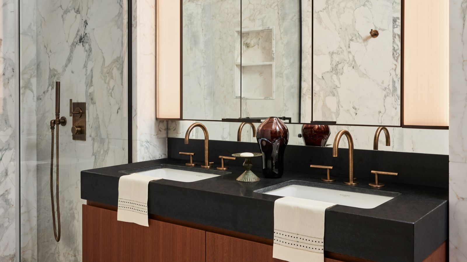 A brown wooden bathroom vanity with a black stone worktop. Two inset sinks with brushed brass faucets and mirrors above. A glass shower with matching showerhead in the background. 