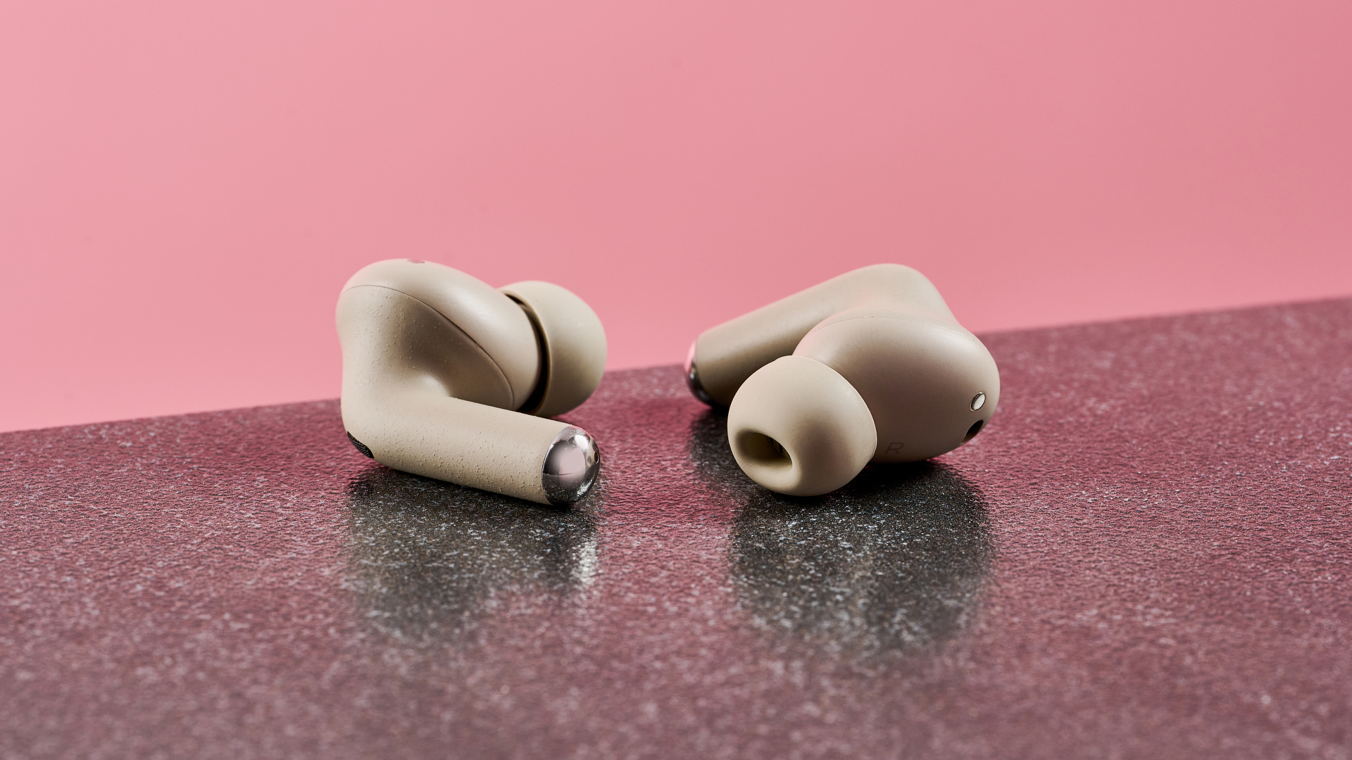 Both Motorola Moto Buds Plus earbuds, one with the silicone tip facing the camera, the other facing backward. They are photographed on a dark surface with a pink background.
