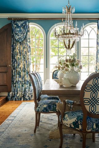 dining room with bright blue walls arched windows wooden table and upholstered blue and white floral patterned chairs and chandelier