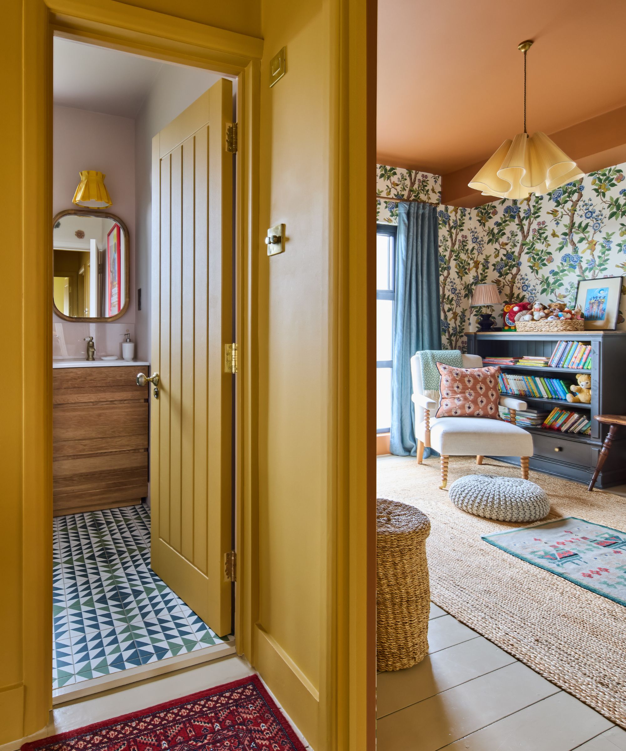 Hallway painted in a warm mustard yellow leading to a floral wallpapered playroom, with a glimpse of a bathroom featuring geometric floor tiles and a wood vanity.