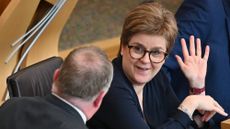Nicola Sturgeon sat at a desk, with one hand up