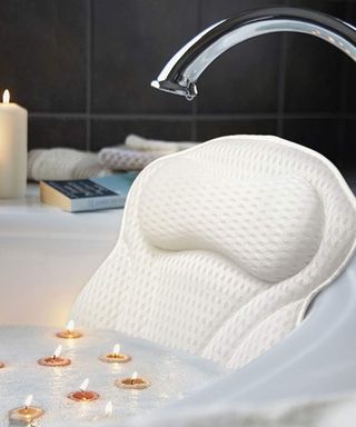 A white bath with a white mesh bath pillow in it, water with bubbles and candles floating on it, a silver faucet above it, and black tiles on the wall