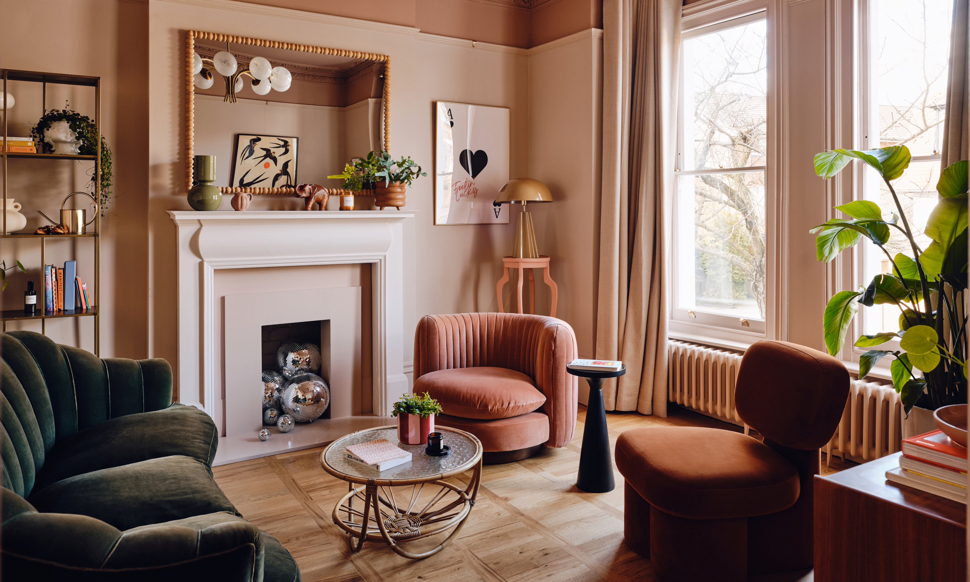 Pink living room with dark green sofa, coral and rust orange armchairs, bamboo and glass coffee table and bamboo mirror over white fireplace