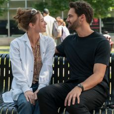 A woman in a lab coat (Lili Reinhart as Olive Smith) and a man in all-black (Tom Bateman as Dr. Adam Carlsen) sit on a park bench, in a still from 'The Love Hypothesis.'