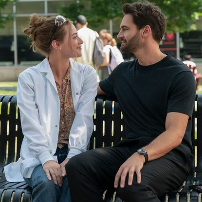 A woman in a lab coat (Lili Reinhart as Olive Smith) and a man in all-black (Tom Bateman as Dr. Adam Carlsen) sit on a park bench, in a still from 'The Love Hypothesis.'