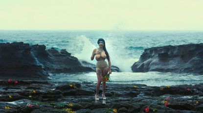 Person in drag standing on rocks, waves crashing behind them