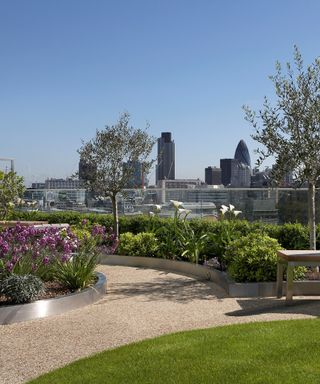 Curved rooftop garden ideas with lawn, gravel and round flowerbeds in front of glass balustrades with city skyscraper views.
