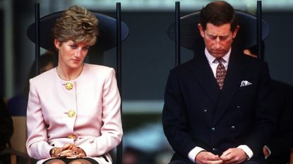 Princess Diana sitting in a chair wearing a pink suit and Prince Charles sitting next to her wearing a dark suit and red tie looking sad