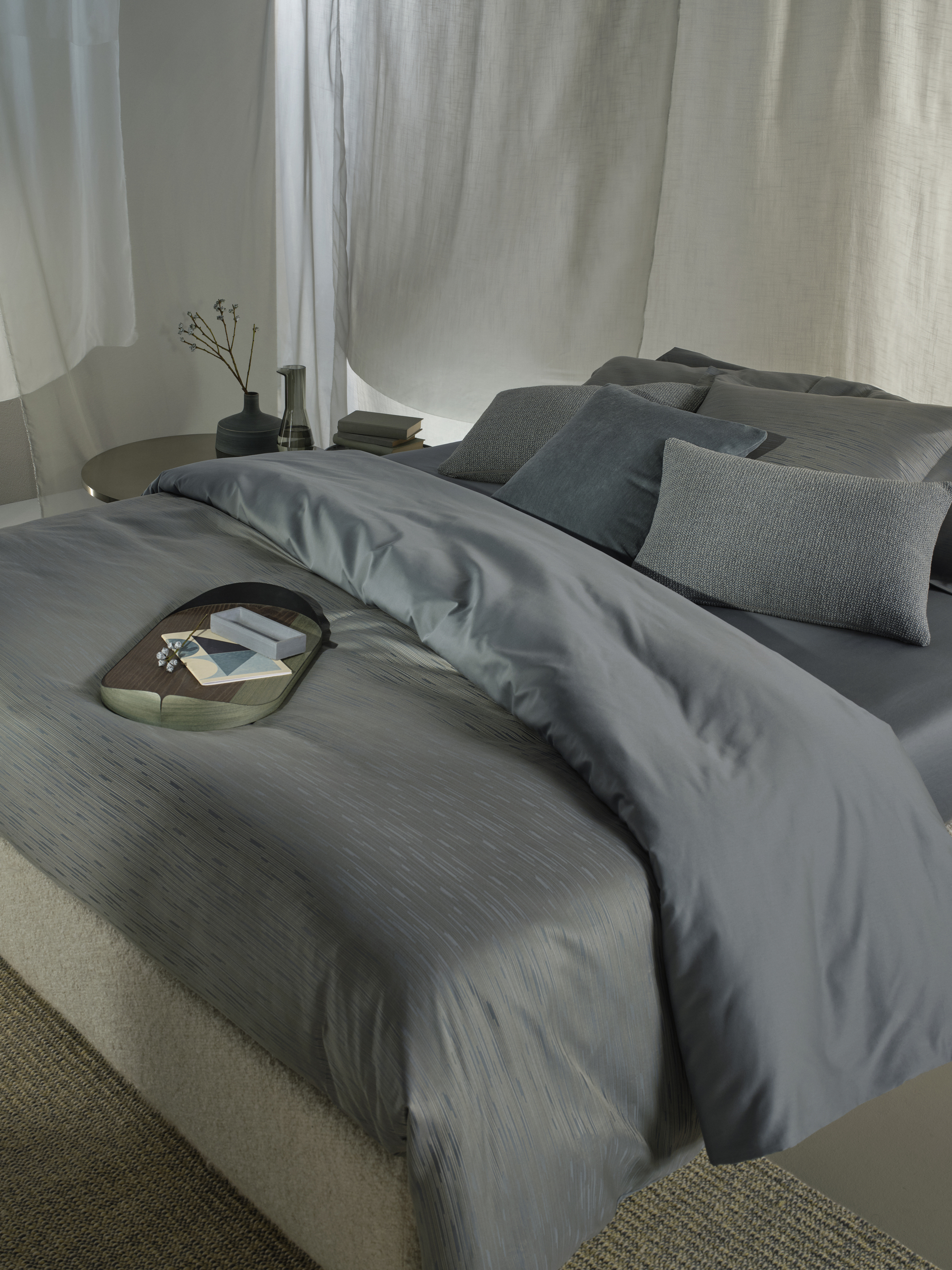 Image of a luxury bed that has dusty blue and stone gray bedding. There is a tray on the end of the bed and curtains on the walls around it.