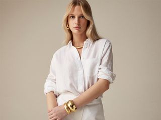 Model wears white button down top tucked into white shorts and gold jewelry.