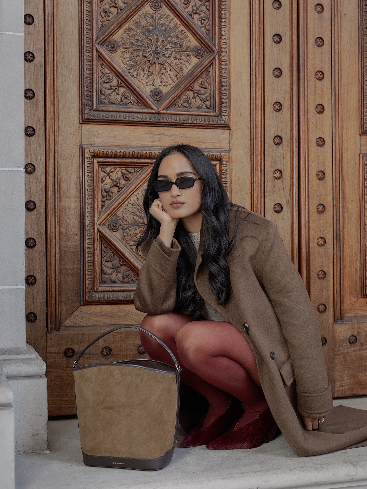 An image of @divrav wearing burgundy tights with a suede bag and a light brown coat.