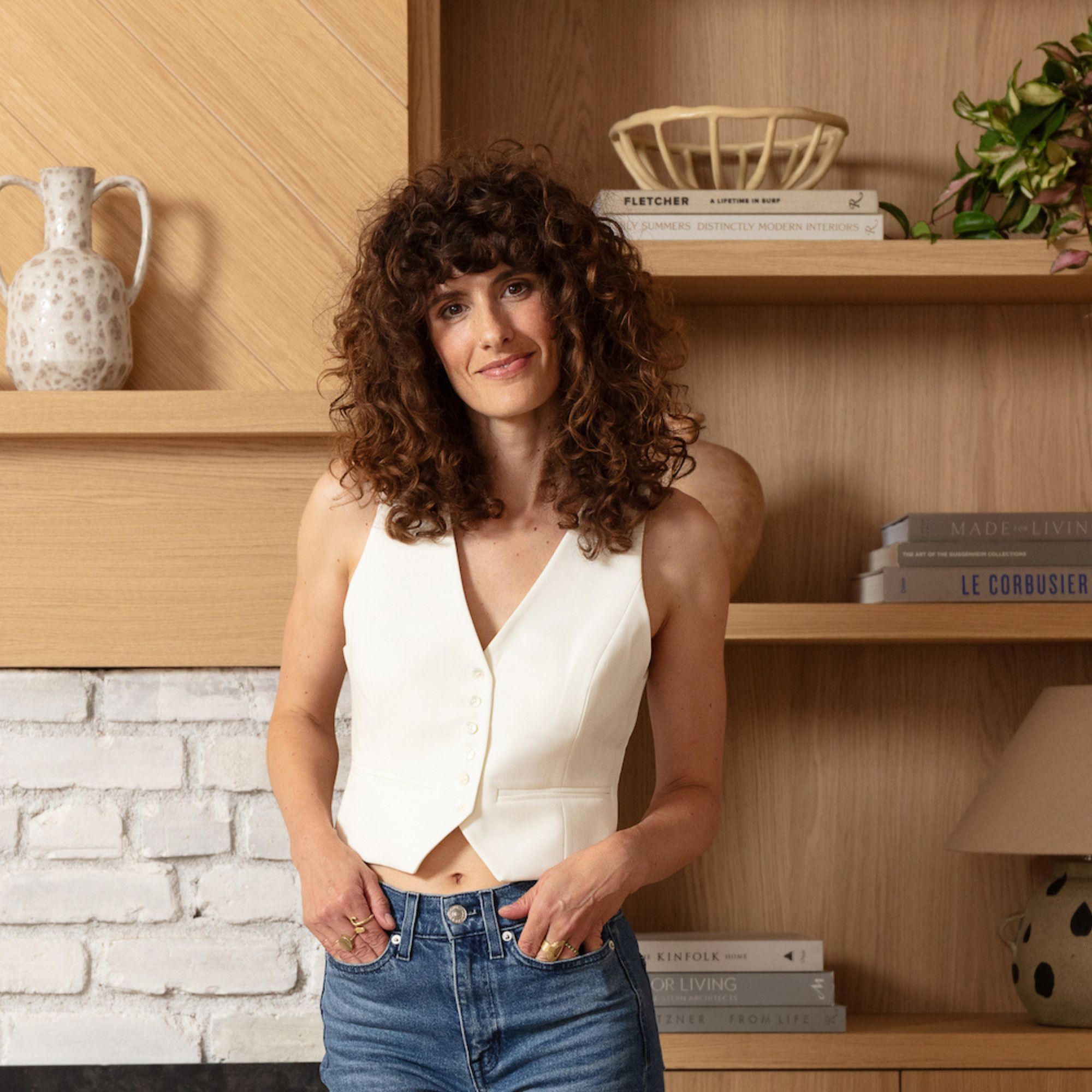 Kara Piepmeyer, a woman with brown curly hair wearing a white waistcoat and jeans, standing in front of wooden shelves with decor on