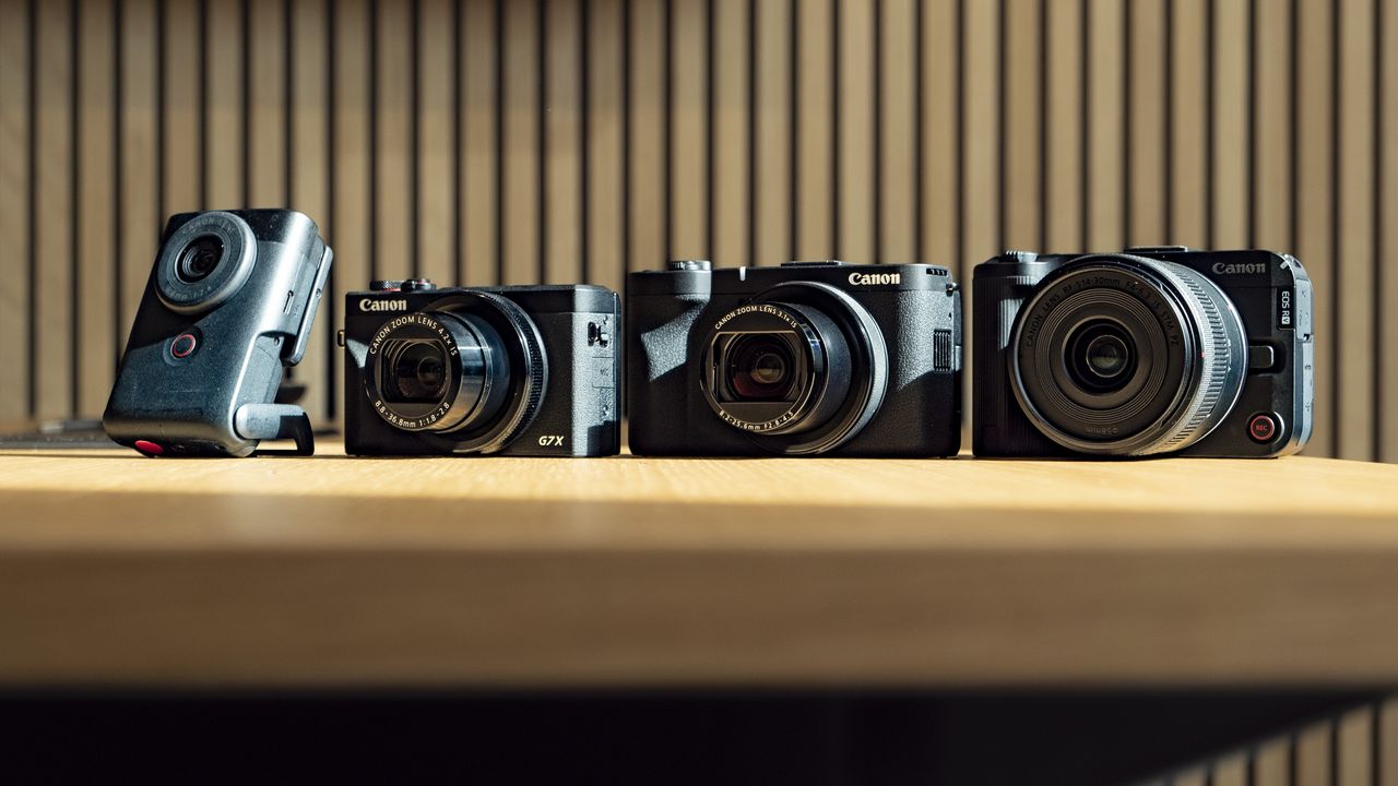 Canon PowerShot V1 compact vlogging camera on a wooden desk alongside the PowerShot G7X Mark III, PowerShot V10 and EOS R50 V