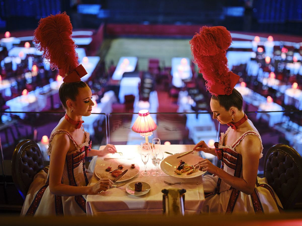 Two dancer women with costumes and a red feather head dine at a romantic table in a lavish setting, featuring dim lighting and exquisite dishes