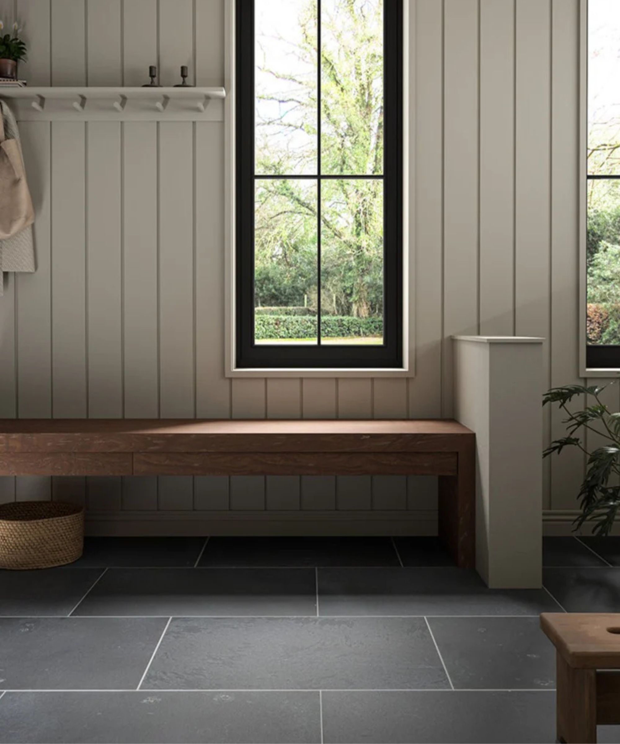 Modern mudroom with vertical panel walls, a long wooden bench, black framed window, and dark slate tile flooring. A woven basket sits beneath the bench, and greenery is visible through the window.