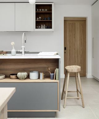 A modern kitchen with a grey and wood panelled island with open shelving, white handleless cabinets, wooden door and stool