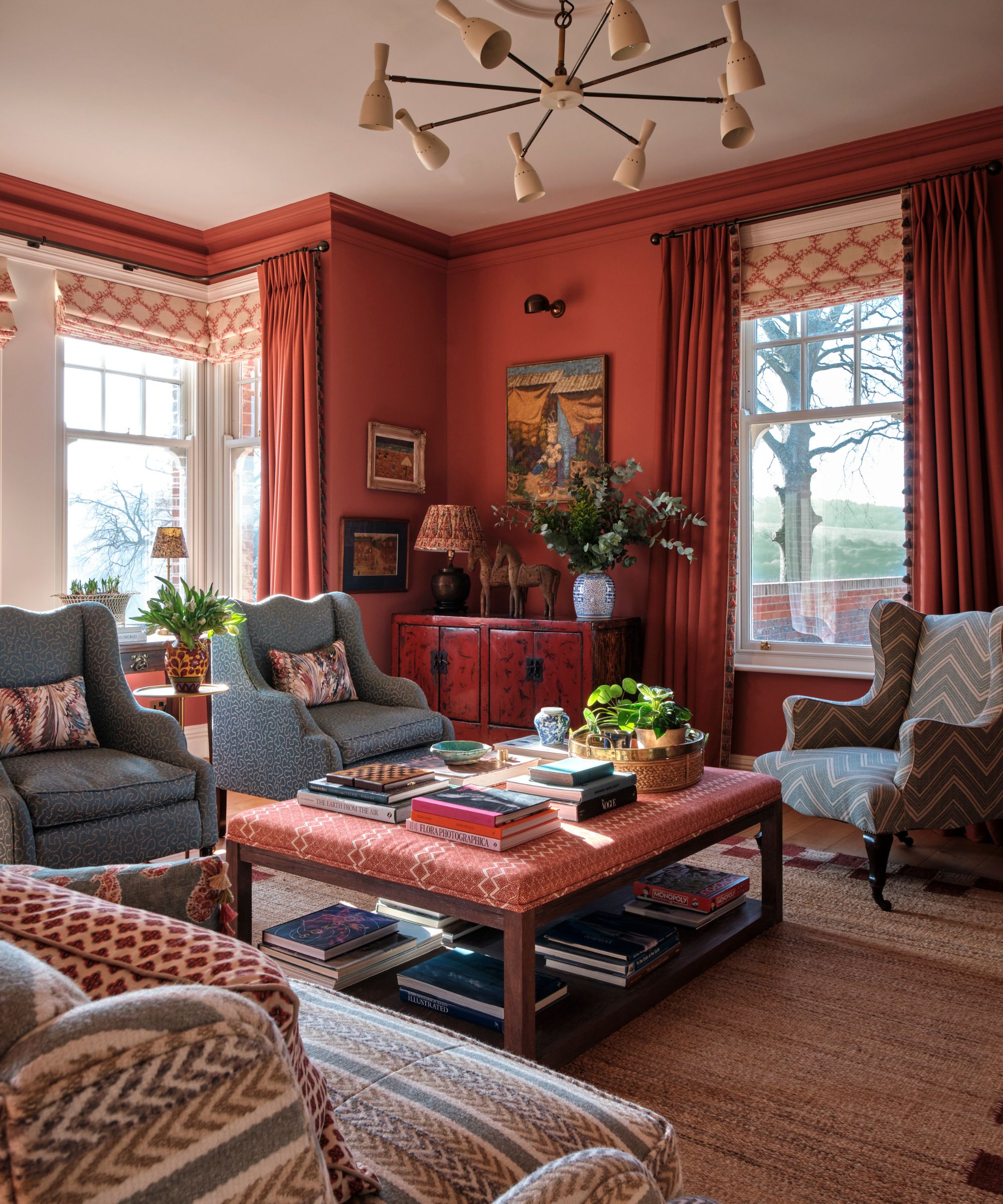Maximalist living room with earthy red walls, upholstered coffee table with books and ceramics, blue armchairs, striped armchair and sofa, midcentury pendant light and rattan carpet
