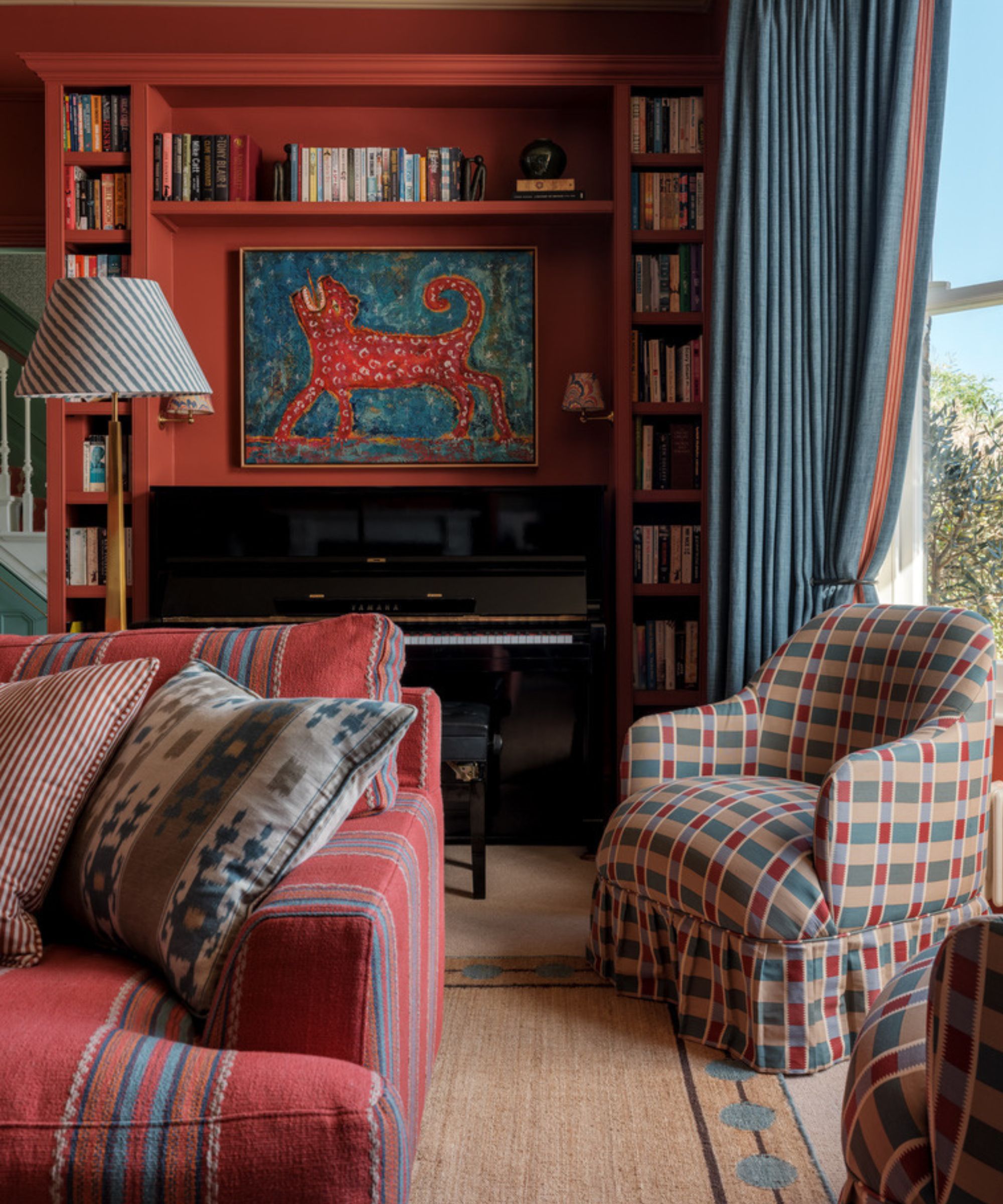 Living room painted red with blue heavy drapes, large bay window, plaid skirted armchairs, a red striped sofa, a jute rug, and a built-in bookcase with a piano and a painting of a leopard beneath.