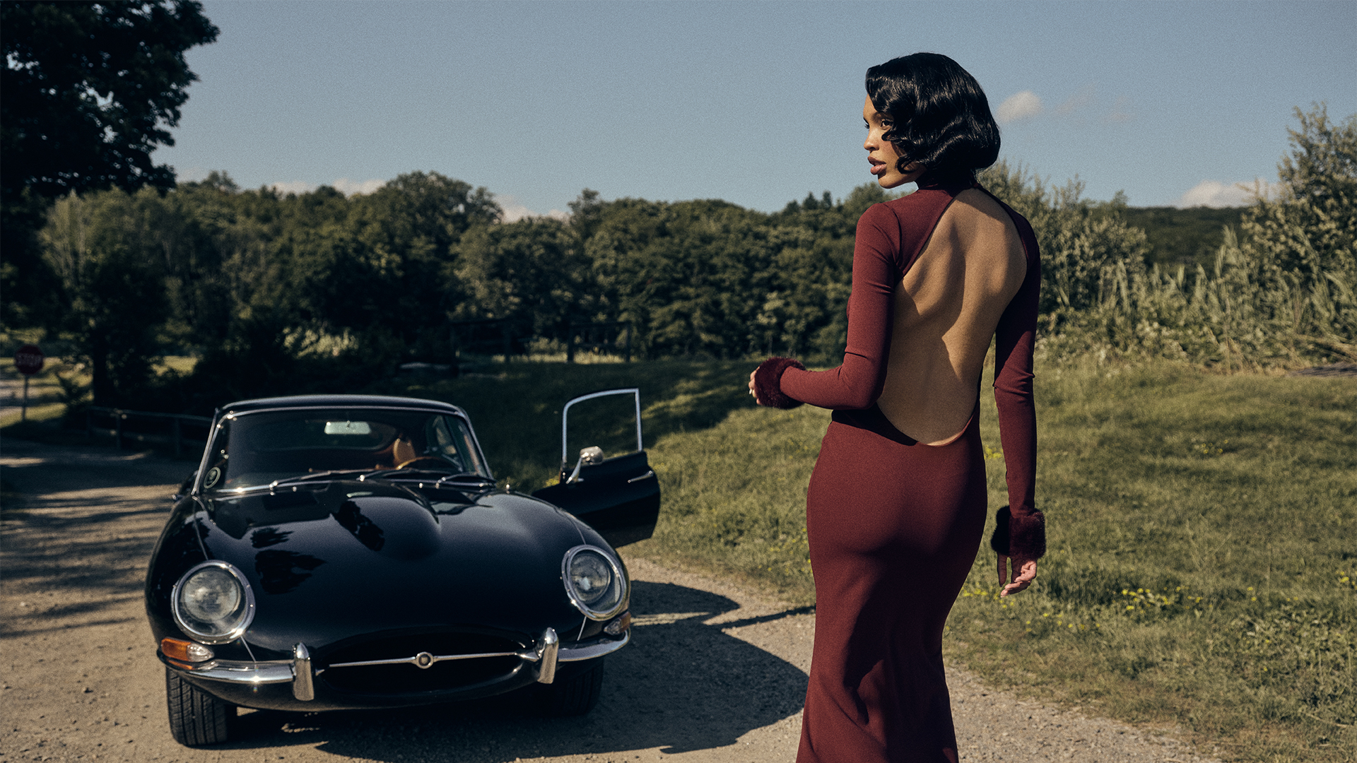 Nara wears read backless dress with fur wrist cuffs while walking towards a black vintage-looking vehicle surrounded by fields