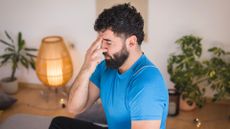 man wearing a blue tshirt, eyes closed, with one hand and finger over his nose, covering one nostril doing alternate nostril breathing. he's indoors with a warm light lamp and plants behind him.