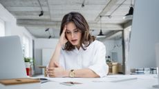 A businesswoman sat at her desk looking sad, with her head in her hand.