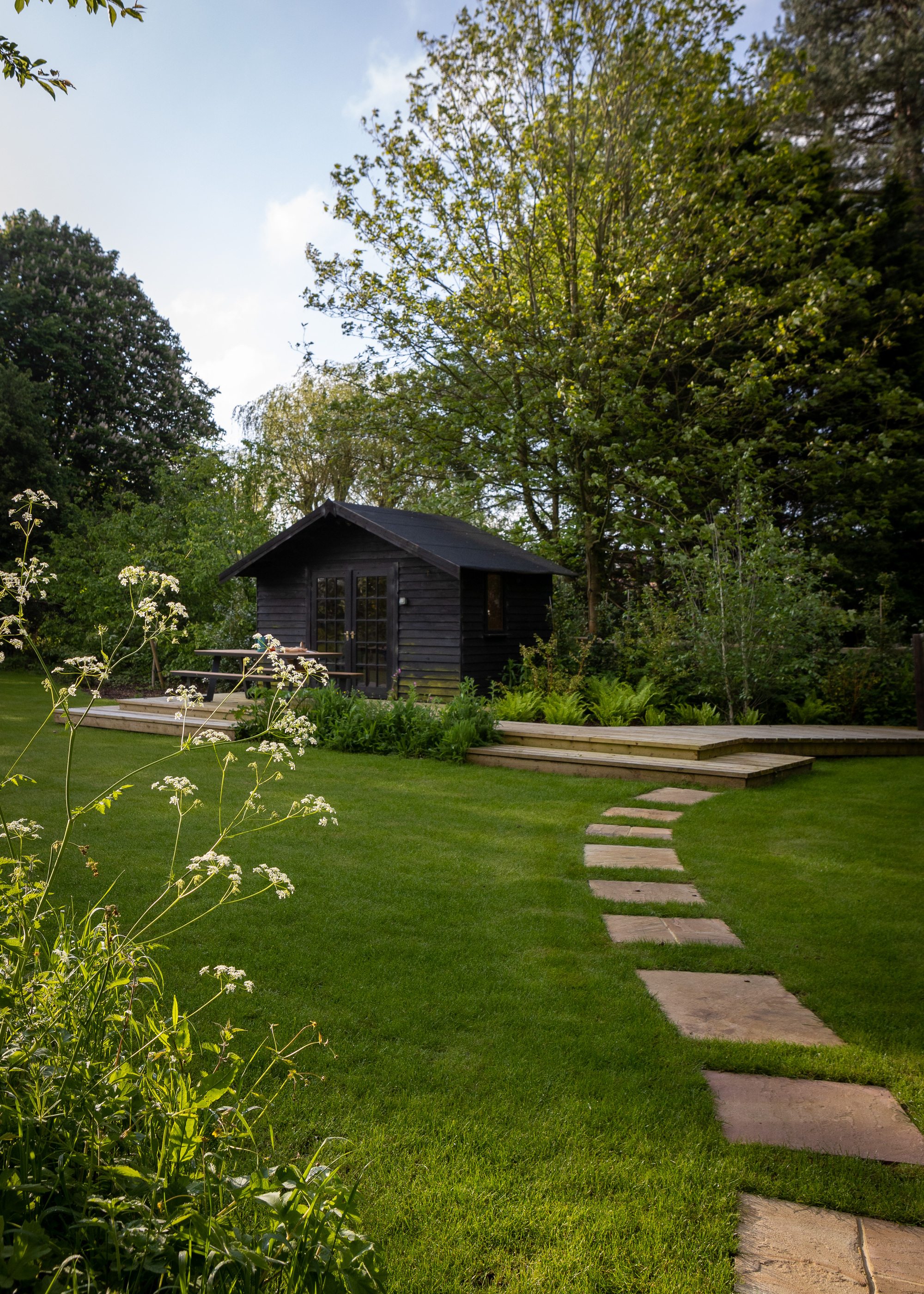 A garden with a large lawn, a paving stone path, and a garden shed painted black on a deck platform