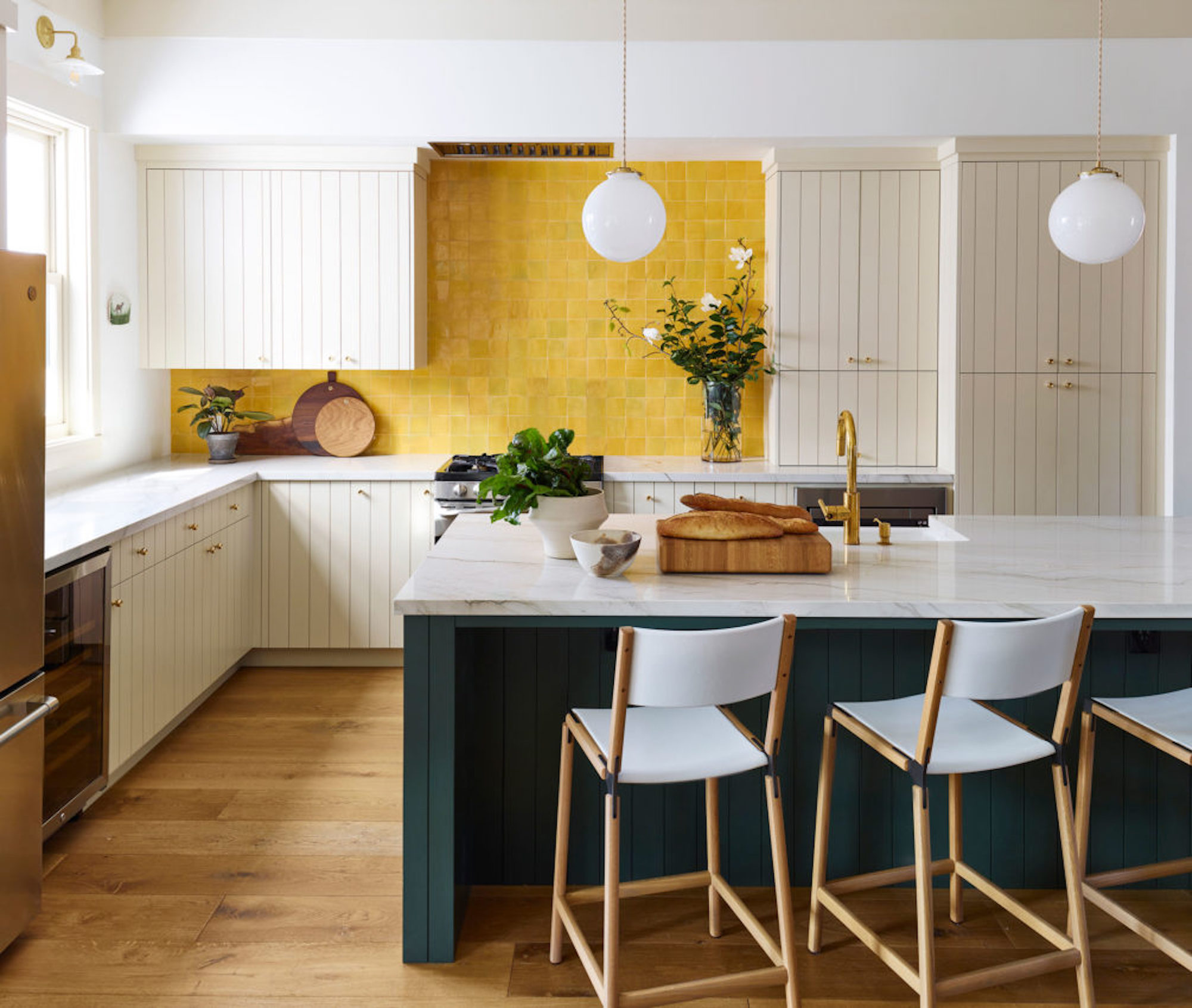 A kitchen with cream coloured, panelled cupboards with sunflower yellow tiles as the backsplash, and a teal kitchen island with marble work tops.