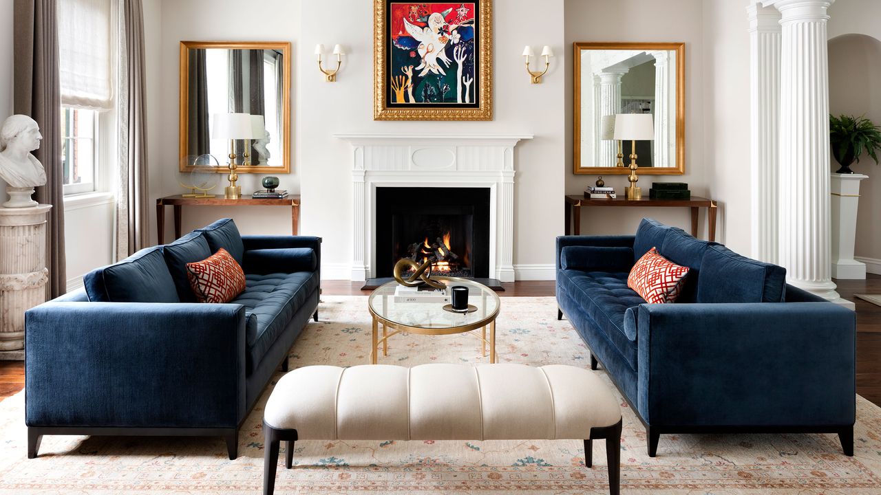 Living room with blue velvet sofas oriental rug fire lit and colorful artwork over mantel mirrors in aloves classical pillar detail and bust statue multipaned windows and white ottoman